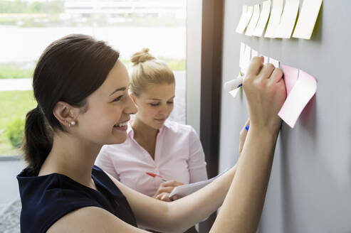 Lächelnde Geschäftsfrau, die auf einen an die Wand geklebten Klebezettel schreibt, während sie mit einem Kollegen im Büro diskutiert - BMOF00426