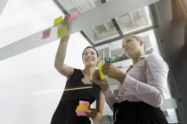 Female colleagues sticking adhesive notes on glass wall while standing in office - BMOF00429