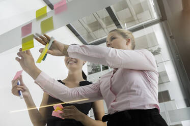 Weibliche Kollegen kleben Haftnotizen an die Glaswand, während sie im Büro diskutieren - BMOF00430