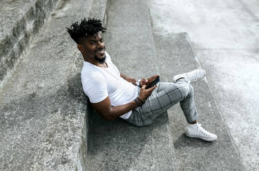 Smiling African man using smart phone while sitting on steps - MGOF04436