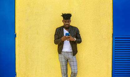 Mittlerer erwachsener Mann in Lederjacke, der ein Mobiltelefon benutzt, während er an einer gelben Wand steht - MGOF04443