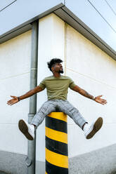 Man with arms outstretched siting on bollard against building in city - MGOF04448