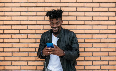 Lächelnder mittelgroßer erwachsener Mann in Lederjacke, der ein Mobiltelefon benutzt, während er an einer Backsteinmauer steht - MGOF04461