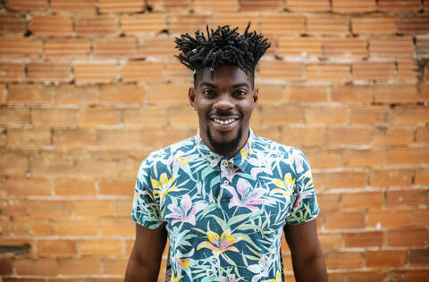 Smiling mid adult man with locs standing against brick wall in city stock photo