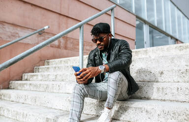 Mid adult man wearing sunglasses using mobile phone while sitting on steps - MGOF04464