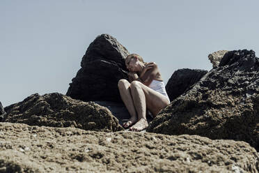 Ältere Frau entspannt sich am felsigen Strand gegen den klaren Himmel an einem sonnigen Tag - ERRF04376