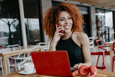 Afro woman with laptop and headphones on table talking over smart phone at sidewalk cafe stock photo