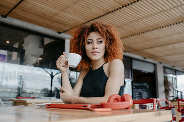 Nachdenkliche junge Frau hält eine Kaffeetasse, während sie in einem Straßencafé sitzt - DSIF00119