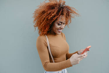 Junge Frau mit lockigem Haar, die ein Mobiltelefon benutzt, während sie an der Wand steht - DSIF00122