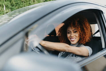 Lächelnde junge Frau mit Afrohaar am Steuer eines Autos, gesehen durch ein Fenster - DSIF00126