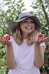 Fröhliche Frau mit Hut und Äpfeln in der Hand auf einem Bauernhof - OGF00590