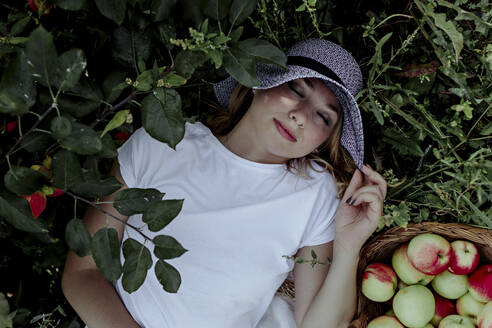 Mid adult woman wearing hat sleeping by apples on land in orchard - OGF00592