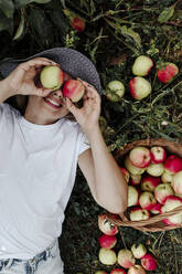 Smiling woman holding apples in front of eyes while lying on land at orchard - OGF00593