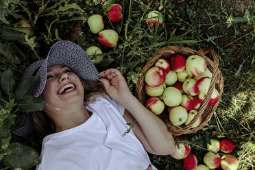Mid adult woman wearing hat laughing while lying by apples on land in orchard - OGF00595