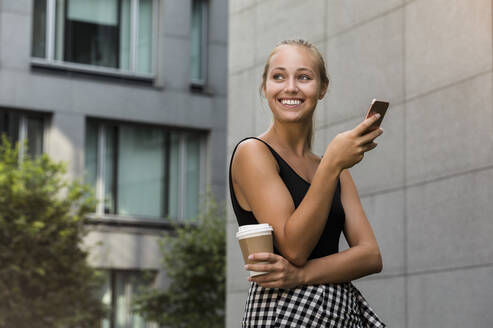 Smiling beautiful woman holding coffee cup using smart phone while standing against building in city - BMOF00442