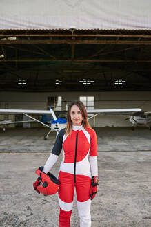 Junge Fallschirmspringerin in einem Hangar mit einem Flugzeug hinter ihr - CAVF89143