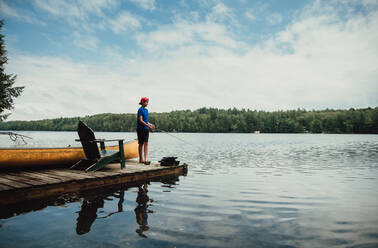 Jugendlicher beim Angeln von einem Steg aus an einem ruhigen See im Sommer. - CAVF89149