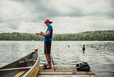 Ein Jugendlicher angelt von einem Steg an einem See, während seine Brüder in der Nähe schwimmen. - CAVF89150