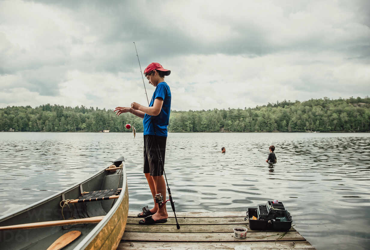 Teen boy fishing from a dock on a lake with brothers swimming nearby. stock  photo