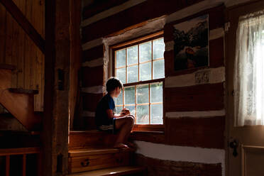Boy sitting on steps of a log cabin cottage looking out through window - CAVF89156