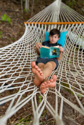 Young boy laying in a woven hammock relaxing and reading a book. - CAVF89162