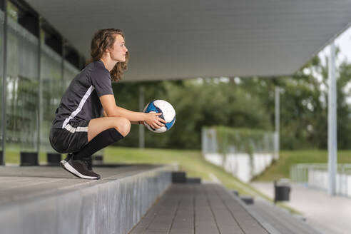 Nachdenkliche Spielerin, die einen Fußball hält, während sie auf den Stufen hockt - STSF02615