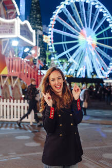 Fröhliche Frau, die vor einem beleuchteten Riesenrad in der Weihnachtszeit steht und gestikuliert - JMPF00399