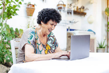 Glückliche ältere Frau, die einen Laptop auf dem Tisch benutzt, während sie im Hof sitzt - PGF00002