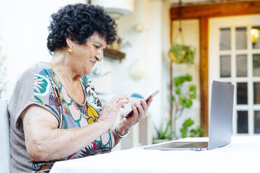 Ältere Frau mit Laptop auf dem Tisch, die ihr Smartphone benutzt, während sie im Hof sitzt - PGF00003