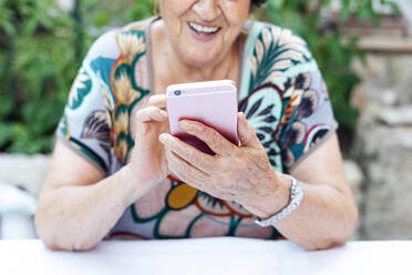 Nahaufnahme einer älteren Frau, die ein Mobiltelefon am Tisch benutzt, während sie im Hof sitzt - PGF00004
