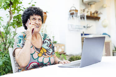 Glückliche ältere Frau mit Laptop auf einem Tisch, die sich über ein Mobiltelefon im Hof unterhält - PGF00007