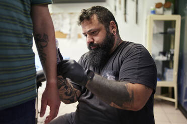 Bearded male artist tattooing on man's hand in studio - SASF00092