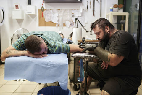 Bärtiger Künstler tätowiert die Hand eines männlichen Kunden auf dem Bett liegend im Studio, lizenzfreies Stockfoto