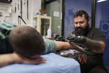 Bearded artist tattooing on mid adult man's hand lying on bed in studio - SASF00095