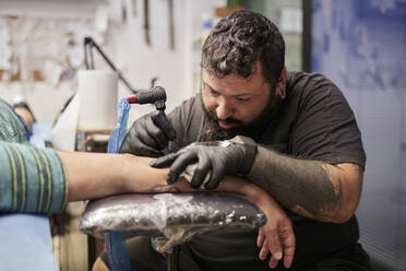 Bearded artist tattooing on male customer's hand in studio - SASF00096