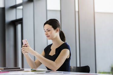Selbstbewusste Geschäftsfrau, die ein Smartphone benutzt, während sie im Büro am Schreibtisch sitzt - BMOF00449