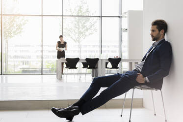 Nachdenklicher Geschäftsmann auf einem Stuhl sitzend, während eine Kollegin im Hintergrund im Büro arbeitet - BMOF00460