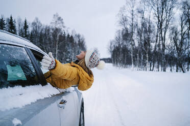 Carefree woman with arms outstretched peeking through car window during winter - DGOF01427