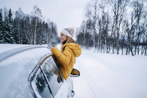 Smiling mature woman sitting on car window against snow covered landscape stock photo