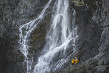 Männlicher Wanderer sitzt auf einem Felsen vor einem Wasserfall, Otscher, Österreich - HMEF01073