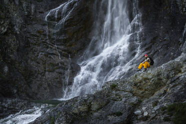 Männlicher Wanderer sitzt auf einem Felsen vor einem Wasserfall, Otscher, Österreich - HMEF01074