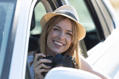 Lächelnde junge Frau, die eine Kamera hält, während sie im Auto sitzt - JSRF01125