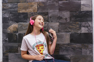 Cheerful girl with headphone listening music while sitting against wall at home - DSIF00128