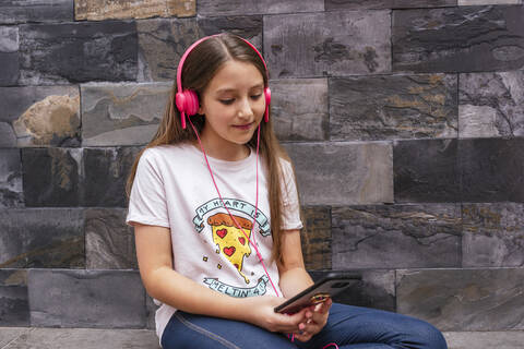 Cute girl with headphones using mobile phone while sitting against wall at home stock photo