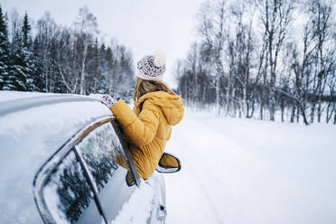 Woman looking at bare trees while leaning out through car window during winter - DGOF01436