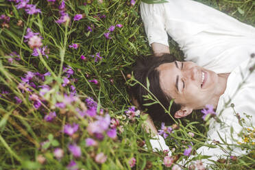 Cheerful woman with eyes closed lying amidst plants on land - EYAF01309