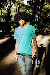 Junger Mann steht mit Kopfhörer in einem öffentlichen Park an einem sonnigen Tag - XLGF00519