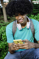 Lächelnder junger Mann, der in einem öffentlichen Park sitzt und mit seinem Smartphone SMS schreibt - XLGF00525