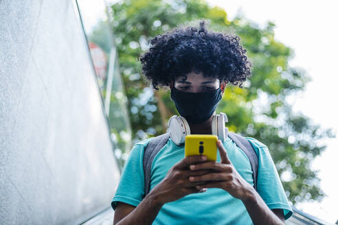 Young man with wearing protective face mask text messaging on smart phone while standing on escalator - XLGF00532