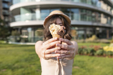 Young woman holding ice cream while standing in public park on sunny day - WPEF03401